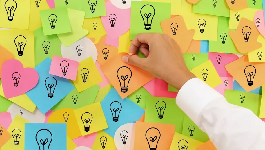 Mão fixando um papel adesivo em painel coberto de notas com ícones de lâmpada